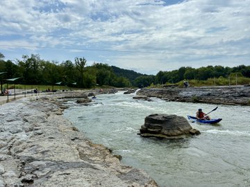 A lone kayaker navigates a rocky, rapid-filled river under a cloudy sky, with a forested shoreline and a small shelter visible in the background.