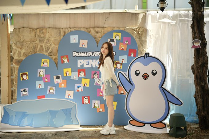 A woman stands between a 'Pengu Playground' photo display wall and a large Pingu penguin mascot cutout at what appears to be a Pengu promotional event.