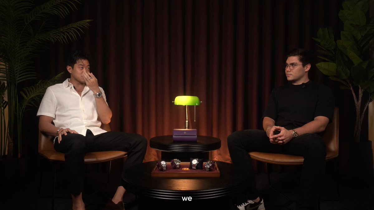 The image shows two men sitting next to each other, both wearing black shirts and pants. They are seated at a table with a green lamp that has a yellow base. The background features brown curtains and a plant. There is also a sign on the wall that reads "Why did we build watchdotfun? Because luxury watches & @solana were always meant to meet, but nobody had built the bridge yet". This suggests that the image might be related to a conversation or event involving watchmakers, watch enthusiasts, or the history of watchmaking.