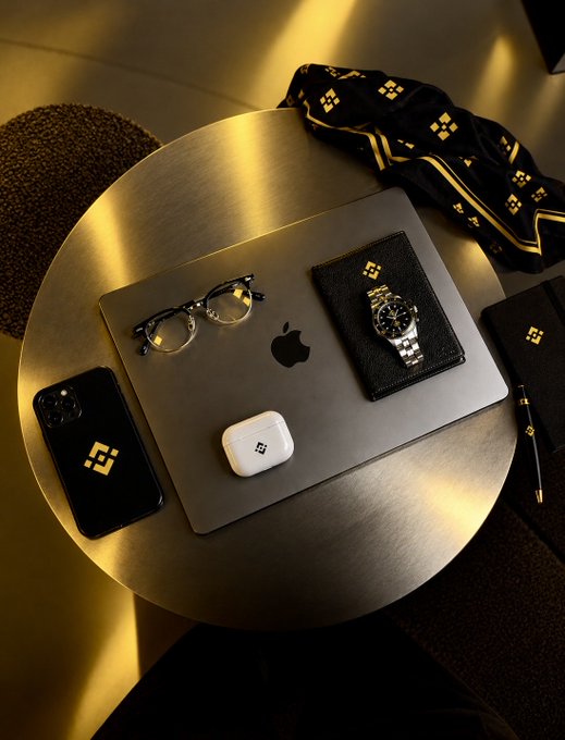 A flat lay photograph displays a golden 'BNB' logo on various items including a phone case, wallet, AirPods case, and scarf, arranged around a laptop on a round table.