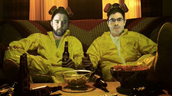 Scene resembling a 'Breaking Bad' parody, featuring two men in yellow hazmat suits and gas masks sitting on a couch surrounded by snacks and beer bottles.