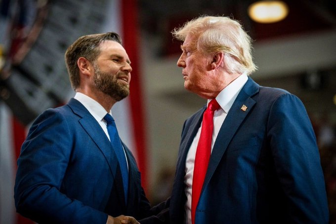 JD Vance and Donald Trump shaking hands and looking at each other, both wearing dark suits with blue and red ties respectively.