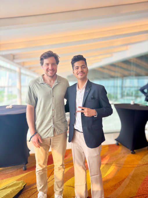 Two young men pose together indoors; one wears a green shirt and beige pants, while the other wears a dark blazer over a white shirt and points toward the camera.