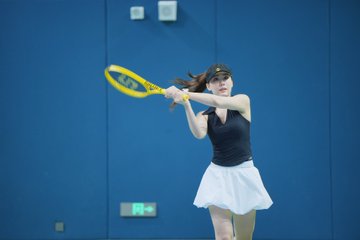 The image features a woman dressed in black, holding a tennis racket and swinging it at an incoming ball during a match. She appears focused and determined as she plays. The background of the image shows a blue wall with a green exit sign on it.