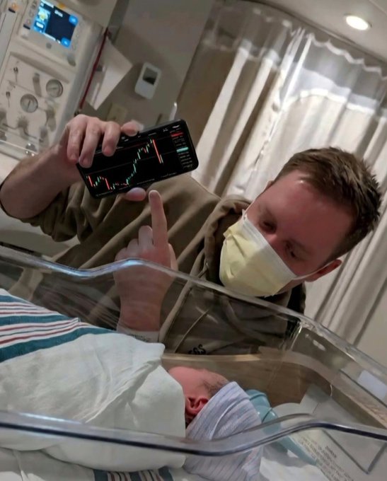 A man wearing a face mask sits in a hospital setting next to a newborn in a bassinet, holding up a smartphone displaying a financial chart.