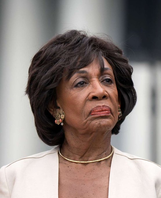 Maxine Waters, wearing a white blazer and ornate earrings, appears in a close-up portrait with a serious expression. The image accompanies her recent statement criticizing Congress for considering cryptocurrency legislation while the President reportedly profits from the industry.