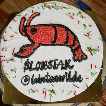 A birthday cake decorated with red and green icing featuring a red cartoon creature design, labeled '$LOBSTAK' and '@lohstawilde'.