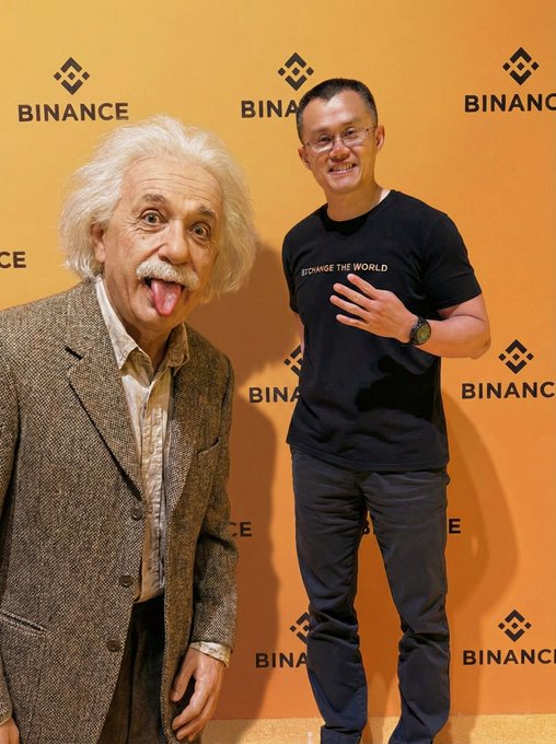A man wearing a 'CHANGE THE WORLD' t-shirt and a mask of Albert Einstein on his hand stands next to a wax figure of Einstein in front of a Binance backdrop.