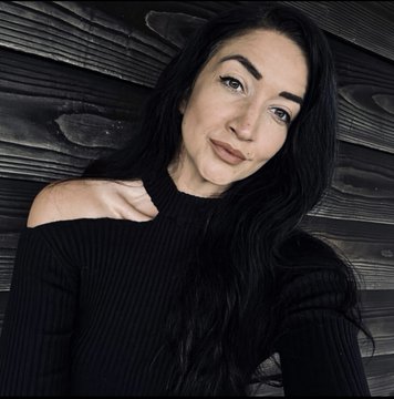 A woman with long dark hair wearing a black off-shoulder sweater poses for a portrait against a dark wooden plank background.