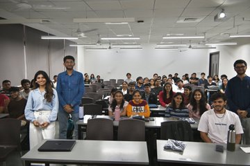 A group photo of students gathered in a bright classroom or lecture hall, with one young woman standing in the front left wearing a white 'GUEST' lanyard, smiling at the camera.