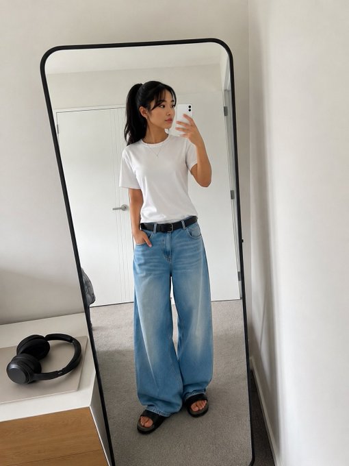 A young woman with a ponytail takes a mirror selfie wearing a white t-shirt, blue jeans, and sandals, with headphones resting on a nearby bench.