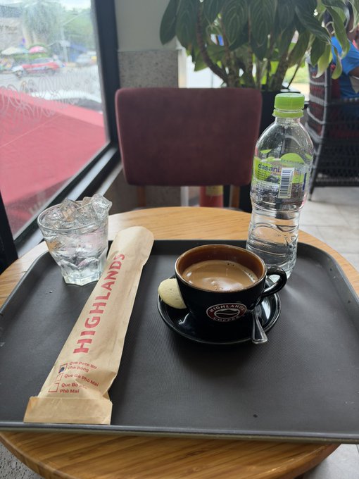 A black tray on a wooden table at a Highlands Coffee shop, containing a cup of coffee with a small cookie, a glass of ice water, a wrapped utensil in a 'HIGHLANDS' branded sleeve, and a bottle of Dasani water.