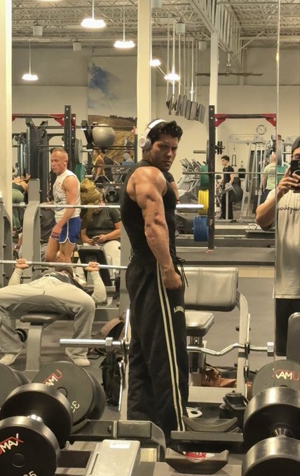 A muscular man in a black tank top and track pants poses in a gym, flexing his arm, with another person taking a photo in the mirror and gym equipment visible around them.