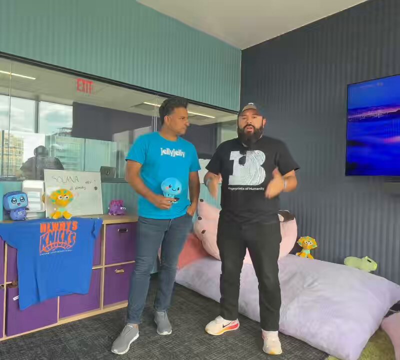 The image features two men standing next to each other, with one of them holding a blue toy. They are positioned near a purple shelf filled with various toys, including stuffed animals such as teddy bears. The background includes a TV screen mounted on the wall above the shelf and another TV screen in the upper right corner of the room.