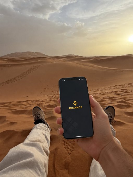 A person sits on the rolling dunes of a vast desert at golden hour, legs stretched out in hiking boots, holding up a smartphone displaying the Binance app logo. The serene, isolated landscape contrasts strikingly with the digital finance platform on screen, capturing the post's message of carrying an entire world of trading possibilities—even in the most remote and quiet places.