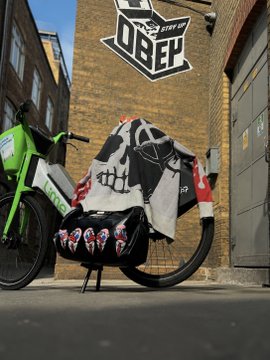 A motorcycle parked on a city street, covered with a black and white skull-themed jacket or cover, in front of a brick wall with an 'OBEY' sticker graffiti. A green and white motorcycle is visible on the left.