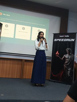 A woman in a white top and blue pants speaking into a microphone at a 'team India SPEEDRUN' event, with a presentation slide visible in the background.