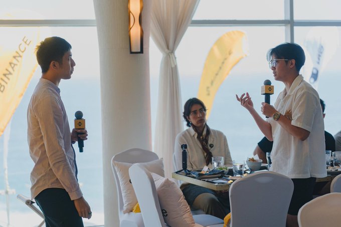 Two men stand holding microphones with the Binance logo at what appears to be a Binance-branded event, with one in a white shirt speaking while another listens, and a seated man with glasses watches from a table in the background.