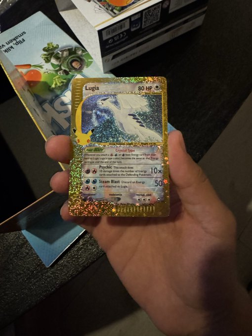 A hand holds a holographic Lugia Pokémon card (80 HP) pulled from a pack, with a Pokémon booster pack and box visible in the background, illustrating the thrill of opening Pokémon packs just like cracking a digital gacha.