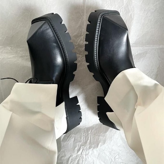 A pair of black platform loafers with chunky lug soles are worn with cream-colored pants, photographed from above against a textured beige surface.