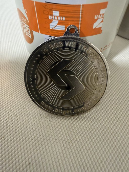 A close-up photo of a metallic commemorative coin/medallion featuring a stylized 'B' lightning bolt logo with the text 'IN BGB WE TRUST' and 'www.bitget.com', resting in front of an orange Bitget cup.