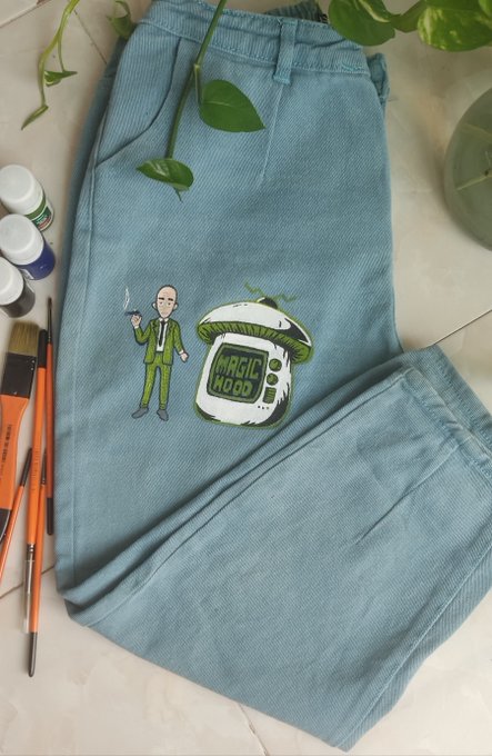 A pair of light blue denim pants laid flat, featuring a hand-painted design of a man in a green suit next to a retro TV labeled 'MAGIC MOOD', with paintbrushes and paint bottles visible nearby.