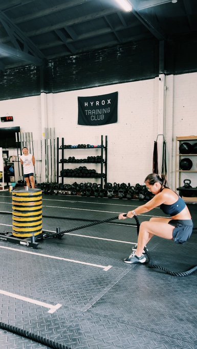 A woman in athletic wear performs battle rope exercises in a gym with a 'HYROX TRAINING CLUB' banner visible in the background, while a trainer observes.