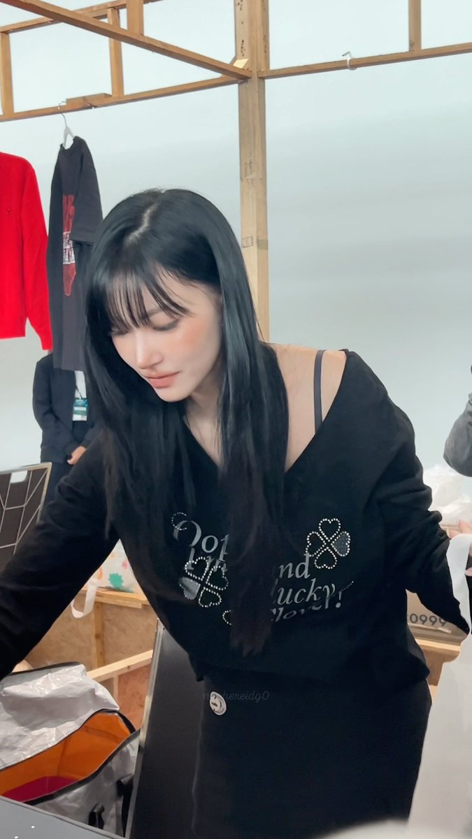 A young woman with long black hair wearing a black top with rhinestone lettering looks downward in what appears to be a retail or studio setting.