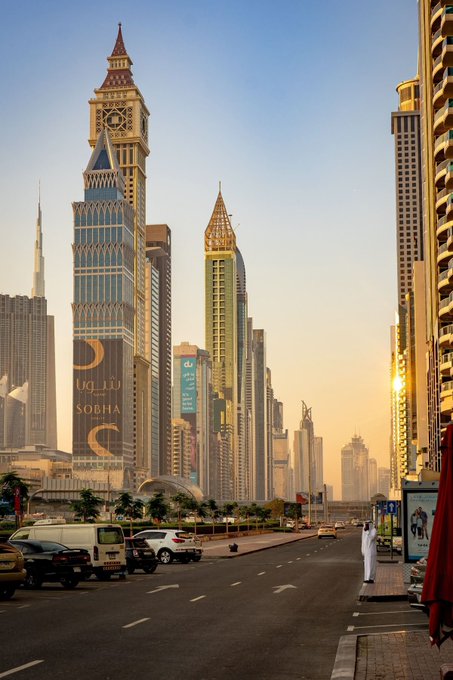 A sunset view of a city skyline with skyscrapers, including one featuring a "SOBHA" advertisement, with vehicles on the street below.
