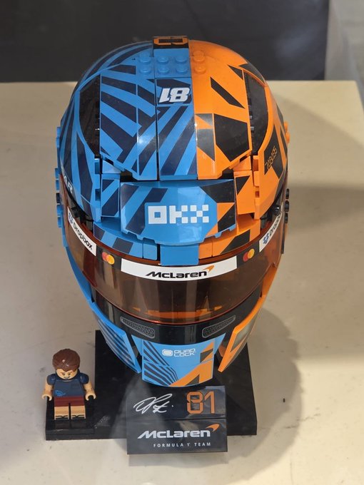 A LEGO McLaren Formula 1 racing helmet model in blue and orange livery, branded with 'OKX', 'McLaren', 'Pure' sponsorships, and the number '81', displayed alongside a small minifigure and a signed nameplate.