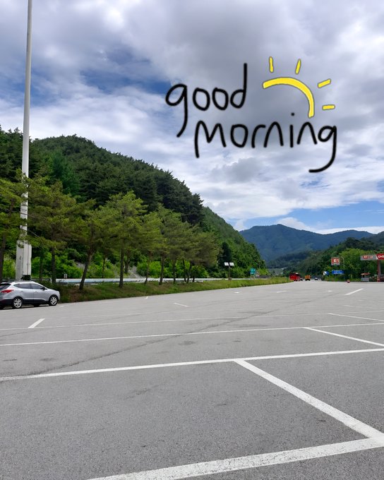 A nearly empty parking lot sits beneath a partly cloudy sky, framed by lush green mountains, with a hand-drawn "good morning" greeting and a smiling sun overlaid on the clouds. The peaceful, early-morning setting mirrors the post's wellness message about starting the day with healthier habits and better sleep.