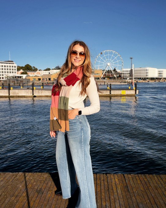 A woman wearing sunglasses, a cream sweater, and a colorful scarf poses on a wooden dock with a Ferris wheel and waterfront buildings in the background.