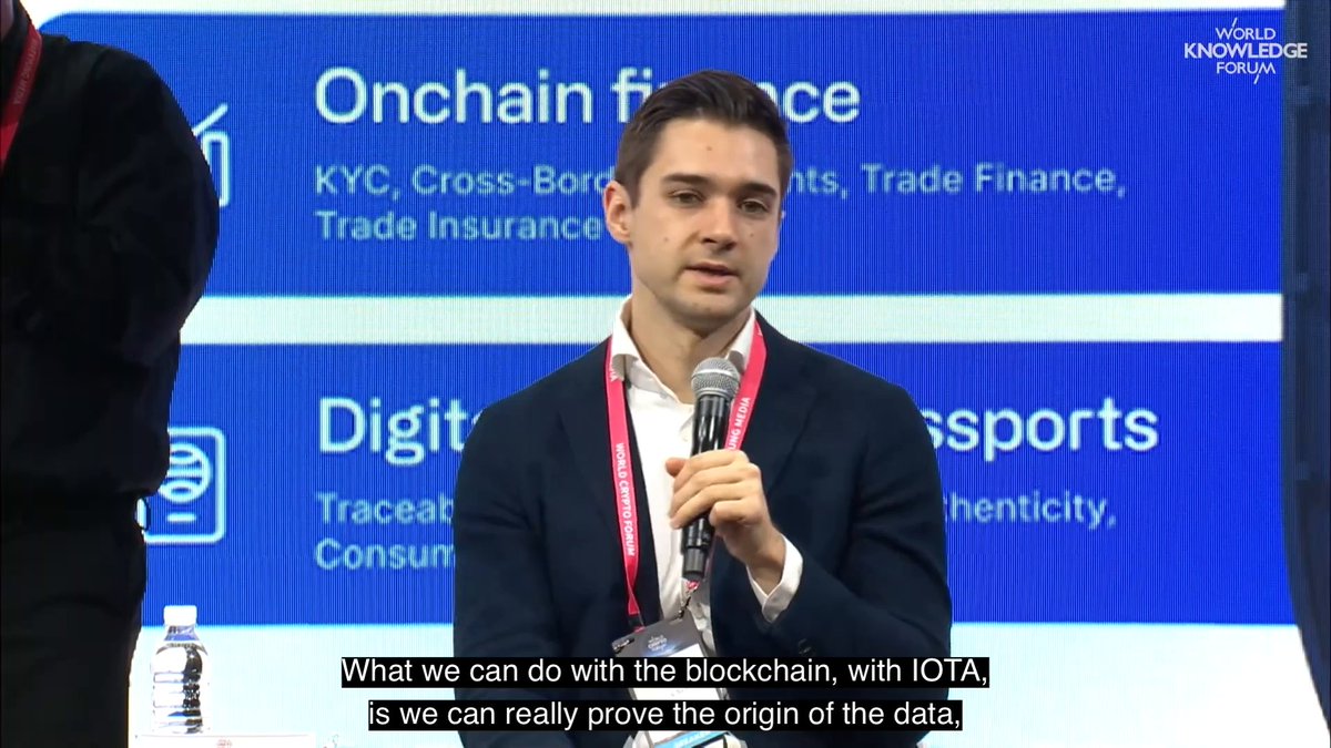 The image shows a man wearing a suit standing behind a microphone, giving a speech or presentation. The background of the image features a blue screen with white text that reads "Onchain Finance", "Digital Trade", and "What we can do with the blockchain is what we really prove the origin of the data". This suggests that the man might be discussing the use of blockchain technology in trade, finance, or other industries. The presence of water bottles on a table near him indicates that this event may have taken place at an event or gathering where attendees are encouraged to stay hydrated and engaged with the speaker's message.