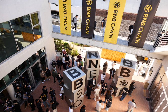 An overhead view of a BNB Chain event, showing attendees gathered in a courtyard with large block letters spelling 'BNB' and 'BUILD' and yellow BNB CHAIN banners hanging above.