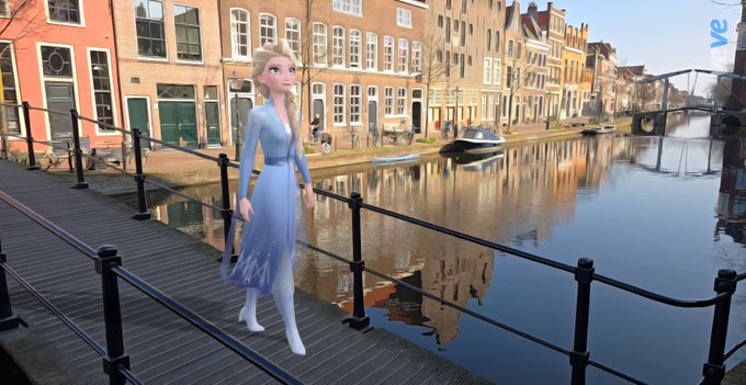 An animated character resembling Elsa from Frozen stands on a wooden bridge overlooking a canal lined with traditional European buildings and boats, with a 've' logo in the top right corner.