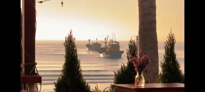The image features a view of boats sailing near the ocean from an outdoor setting with potted plants and trees. There are two boats visible in the scene - one larger boat towards the left side, and another smaller boat closer to the center-right area. The boats appear to be on the water, possibly enjoying their time out on the sea. In addition to the boats, there is a vase with flowers placed near the right edge of the image, adding a touch of color and life to the scene.