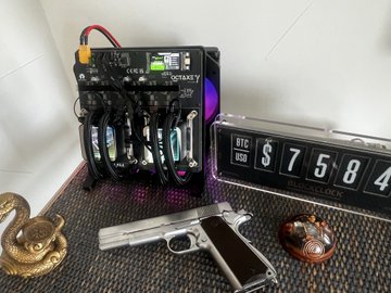 A photograph of cryptocurrency mining equipment, including an ASIC miner and multiple GPUs, alongside a Bitcoin clock displaying '67584' USD, a pistol, and other miscellaneous items on a desk.