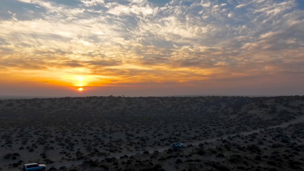 The image shows an aerial view of a desert landscape with a beautiful sunset casting warm hues over the scene. The sun is setting behind the horizon, creating a stunning backdrop for the sky filled with clouds. In the foreground, there are several small vehicles parked on a dirt road that cuts through the desert. One car can be seen in the middle of the frame, while another is located closer to the right side. A person is also visible near one of the vehicles, possibly attending to their vehicle or enjoying the view. The image captures the essence of an open-world adventure and exploration, with a sense of freedom and tranquility.