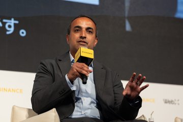 A man in a dark blazer and light blue shirt speaks into a yellow microphone at what appears to be a conference, with partial text 'ई.' visible in the background.
