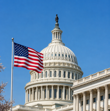 The U.S. Capitol building stands beneath a clear blue sky, with an American flag waving prominently in the foreground—symbolizing the legislative delay of the CLARITY Act, which failed to reach the Senate floor before summer recess as reported by Majority Leader Thune.