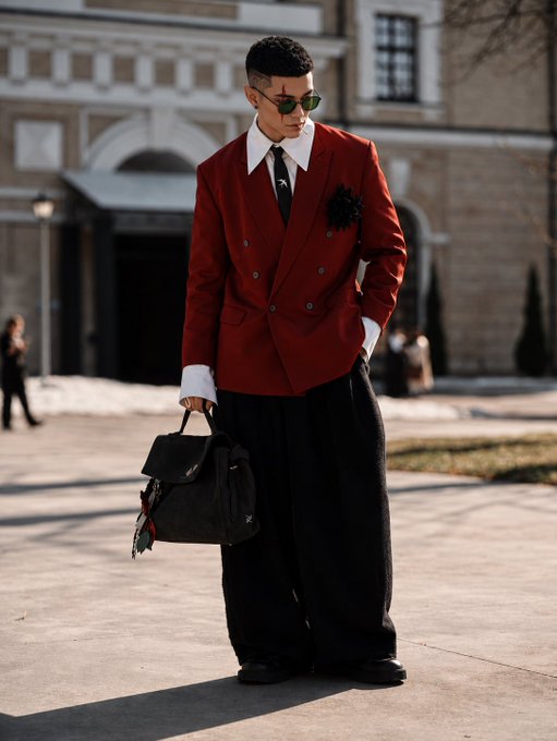 A fashion photo of a man in a red double-breasted blazer, white collared shirt, black tie, and oversized black wide-leg pants, holding a black Louis Vuitton bag while wearing sunglasses outdoors.
