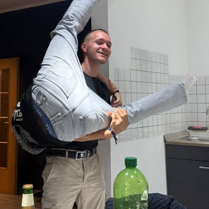 A smiling man in a kitchen lifts another person upside down by their legs, with a large green soda bottle and tiled wall visible in the background.