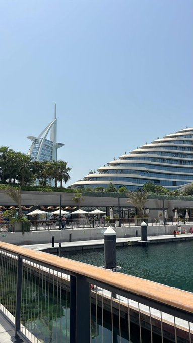 A waterfront view featuring the Burj Al Arab hotel and the Jumeirah Beach Hotel under a clear blue sky, with outdoor dining umbrellas lining the promenade.