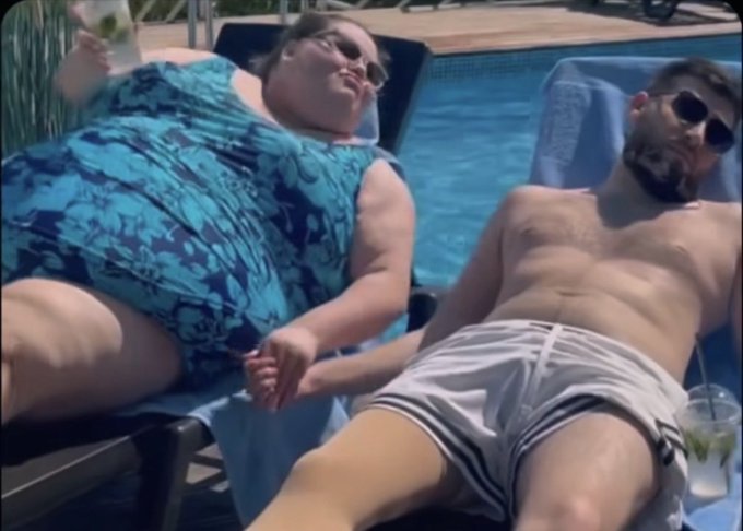 A candid photo of a couple relaxing on pool lounge chairs in swimwear and sunglasses, with the woman holding a drink, enjoying a sunny day by a swimming pool.