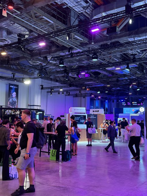 A crowded indoor convention or trade show with purple lighting, featuring attendees and various exhibitor booths.
