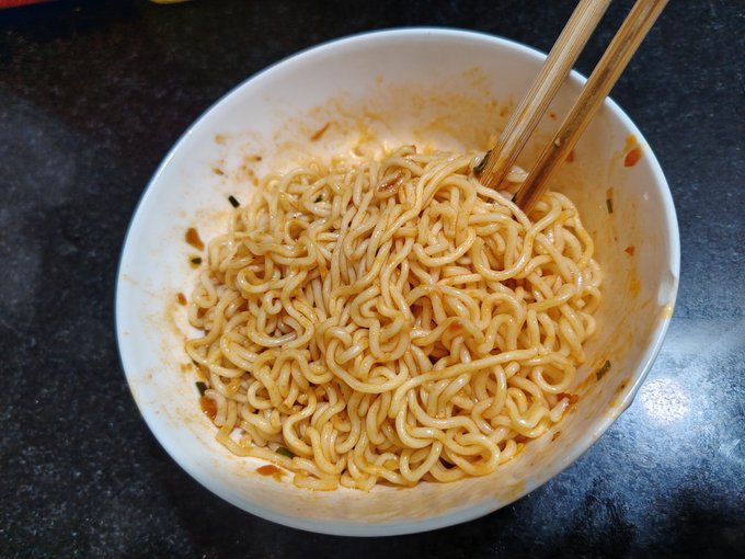 A white bowl filled with cooked noodles coated in a reddish sauce, with a pair of wooden chopsticks resting inside, placed on a dark countertop.