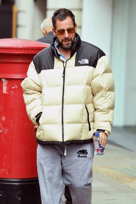 Actor Adam Sandler walking outdoors wearing a cream-colored North Face puffer jacket, sunglasses, grey Roots sweatpants, and holding a water bottle.