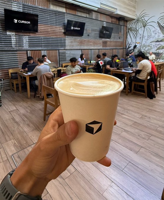 A hand holds a coffee cup branded with the Cursor logo in the foreground of a coworking space where developers work on laptops, with "CURSOR" displayed on wall-mounted monitors in the background. The image visually reinforces the post's message about engaging with AI-focused developer communities like Cursor to grow the Solana ecosystem.