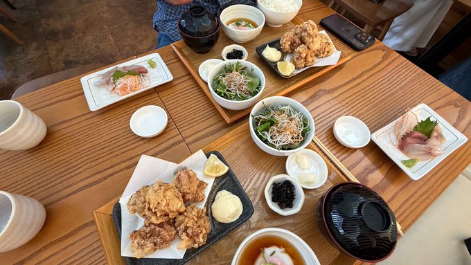 An overhead view of a Japanese meal spread on a wooden table, featuring fried chicken karaage, sashimi, rice bowls, miso soup, and various side dishes.