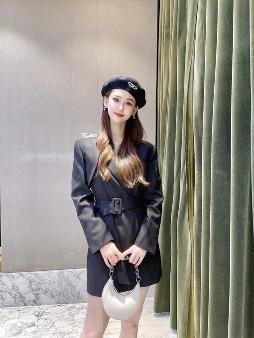 A woman poses wearing a black beret with a logo, a belted dark grey coat dress, and holding a silver crescent-shaped handbag.